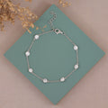 A delicate silver chain bracelet with eight small, round clear crystal or pearl-like beads, displayed on a teal square card over a beige background with dried floral accents.
