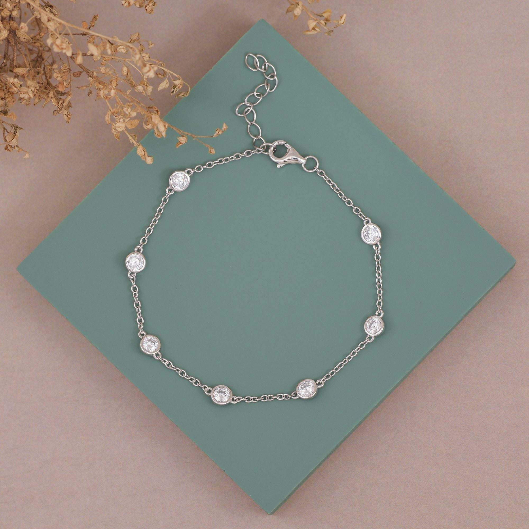 A delicate silver chain bracelet with eight small, round clear crystal or pearl-like beads, displayed on a teal square card over a beige background with dried floral accents.